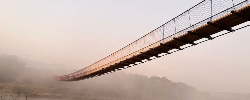 A suspension bridge swoops across a river through the early morning mist