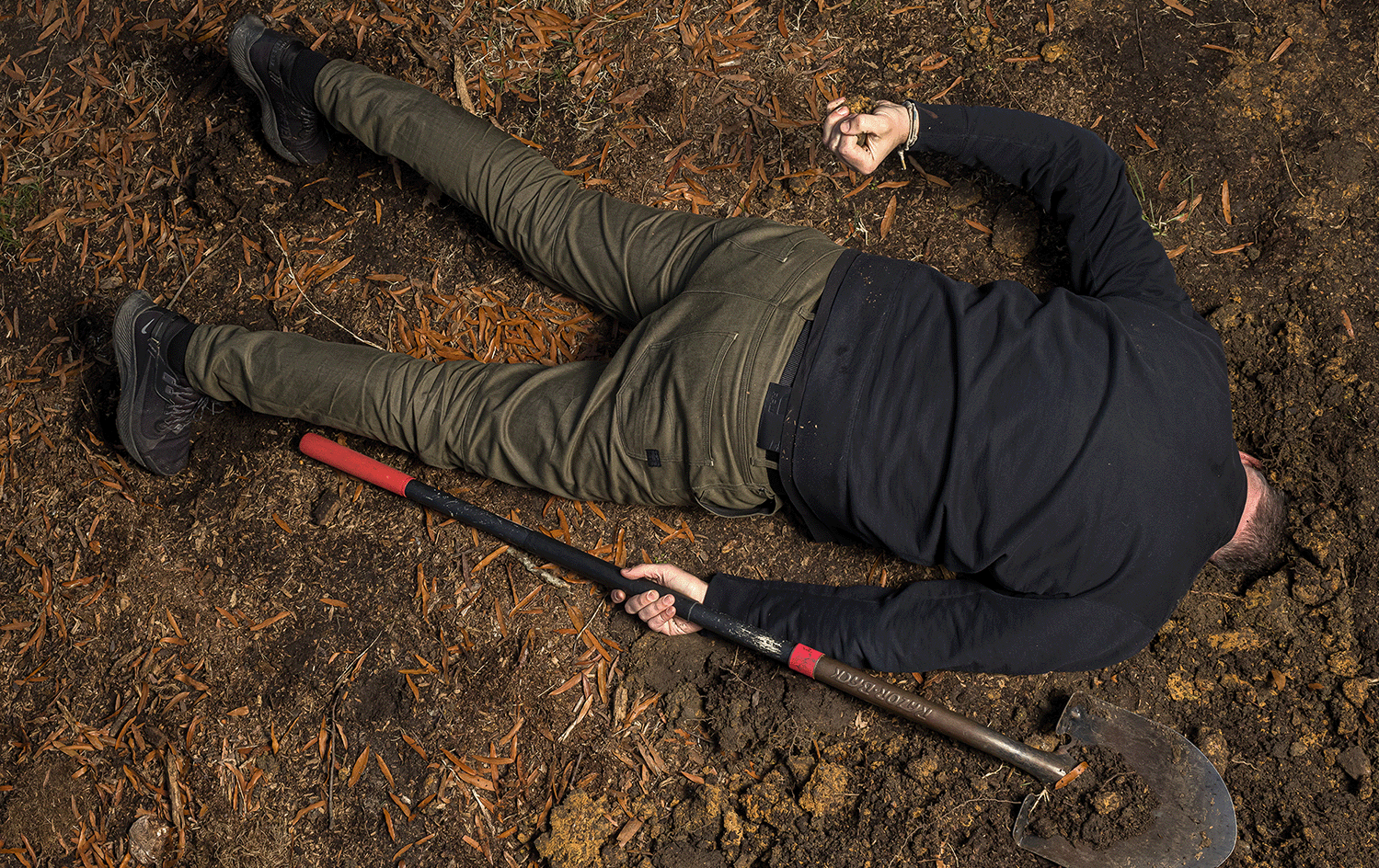 A man lies face down in the ground, with a shovel in one hand and the other clenched around a fistful of dirt.