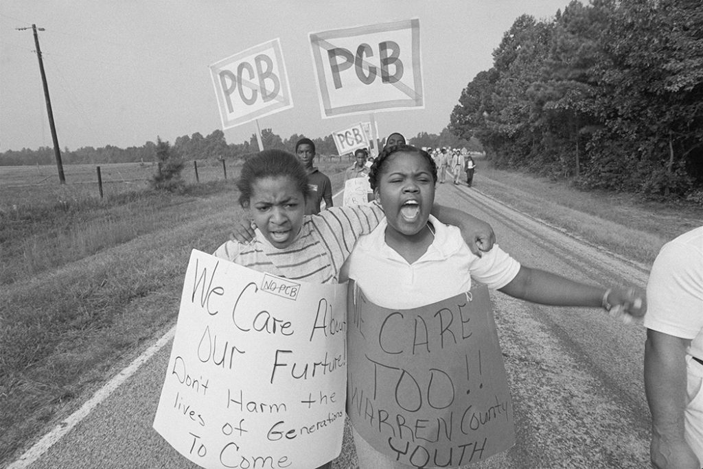 Two girls walk down a rural road holding protest signs as other protestors follow