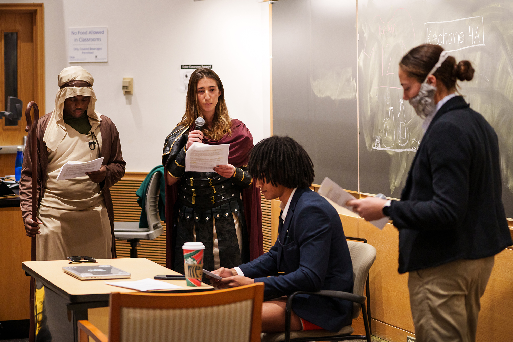 Three students dressed as philosophers and religious figures offer advice to another student in a blazer, sitting at a desk.