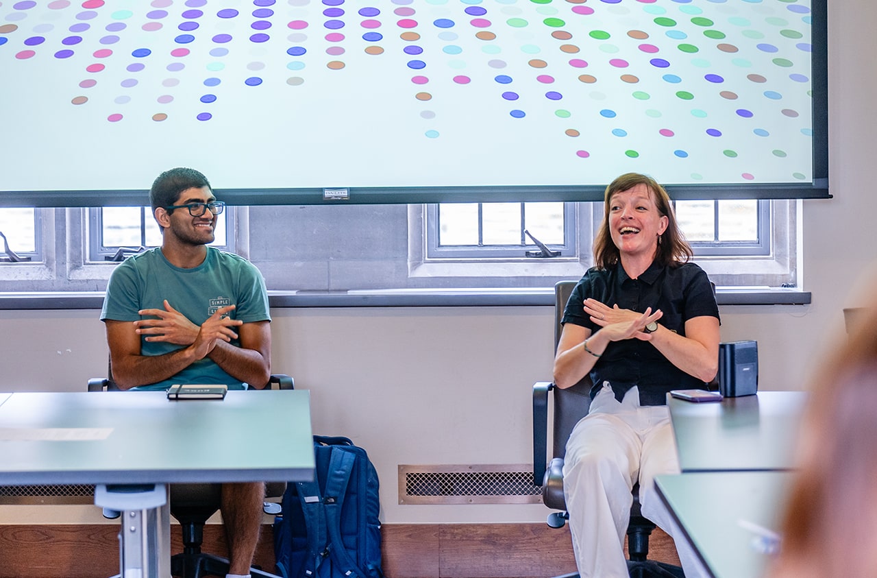 A student and faculty member sitting in front of a classroom laugh as they move their hands in the same motion.