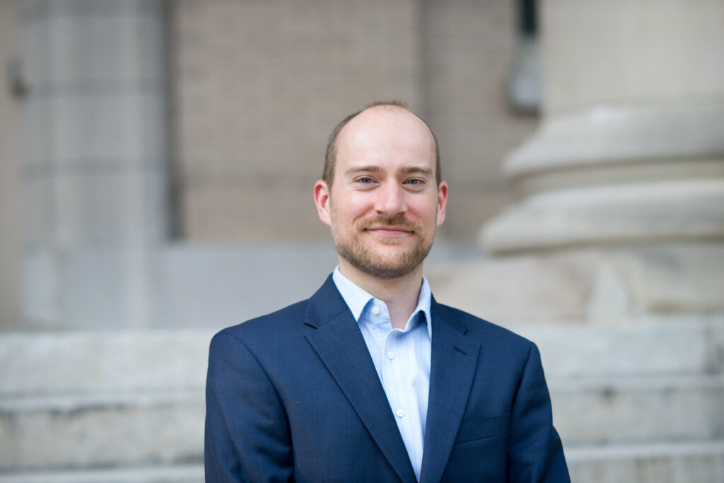 Headshot of a man in a navy blazer standing in front of a building with columns